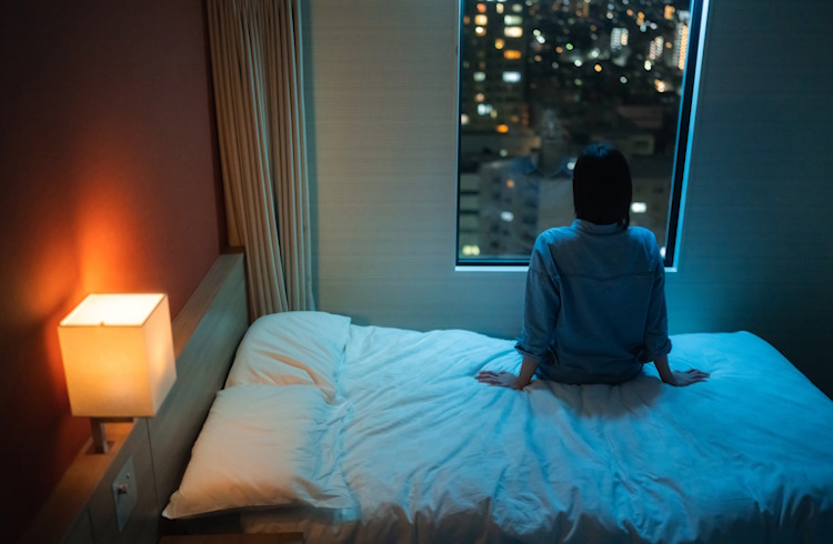 A woman sits in her hotel room in the middle of the night, looking out at the city.