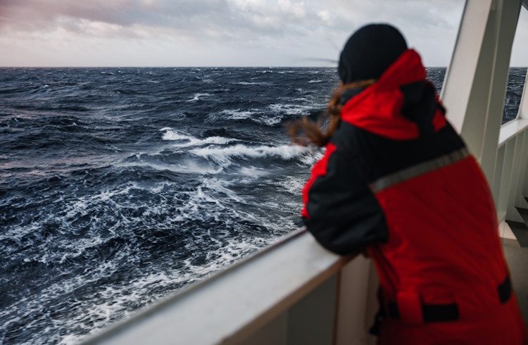 A traveler stands on the deck of an expedition ship looking out over rough seas. 