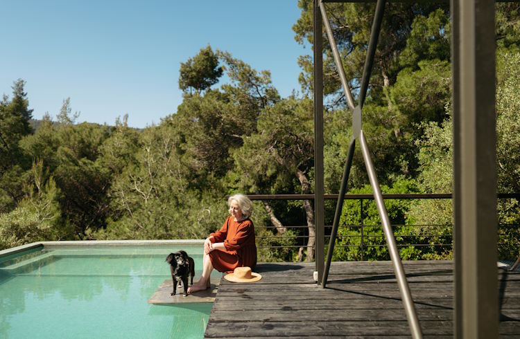 A senior woman sits with her dog at the edge of a swimming pool in Greece.