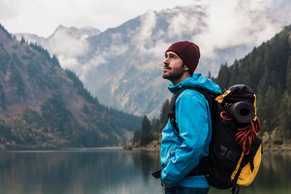 A man hiking in front of a mountain range, he is wearing warm weather gear and a backpack