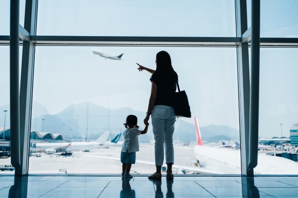 Rear view of young Asian mother holding hands of cute little daughter looking at airplane through window at the airport while waiting for departure.